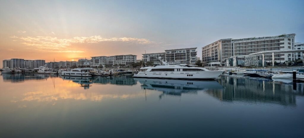 Panoramic view of a marina at sunset, featuring luxury yachts and modern waterfront buildings reflecting in the calm water.