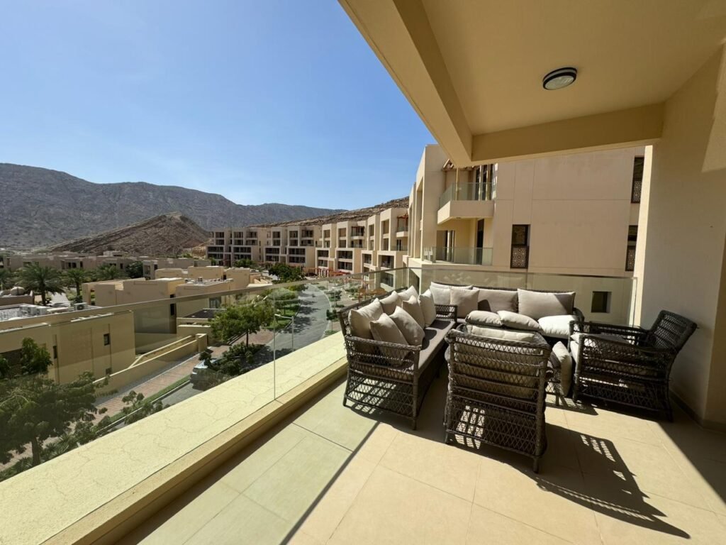 A spacious balcony featuring wicker furniture with cushions, overlooking a scenic view of mountains and nearby buildings under a clear blue sky.