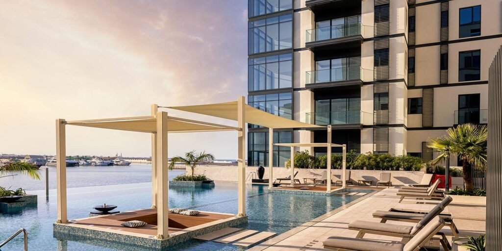 Luxurious swimming pool area with canopies and lounge chairs, overlooking a serene waterfront and modern high-rise building.