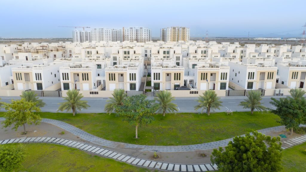 Aerial view of a residential area featuring multiple white houses lined up, with palm trees and green grass in the foreground and modern buildings in the background.